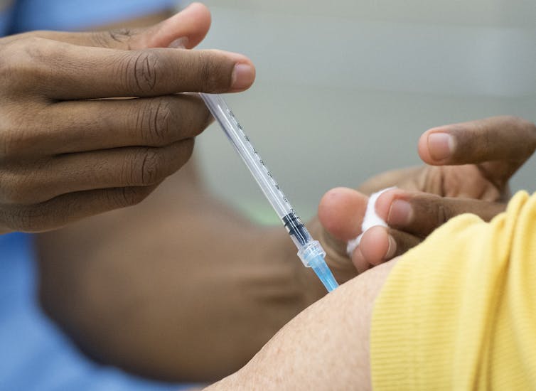 Close-up of a person's shoulder in yellow T-shirt getting an injection in the upper arm.
