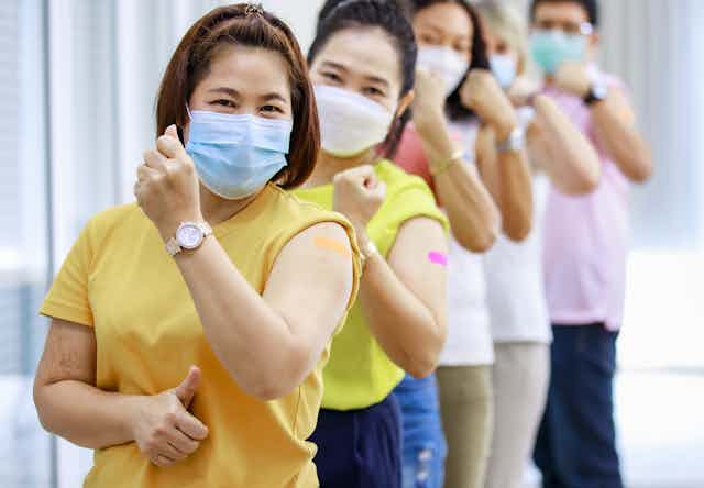 Five people lined up wearing facemasks and flexing their arm with a bandaid on the bicep