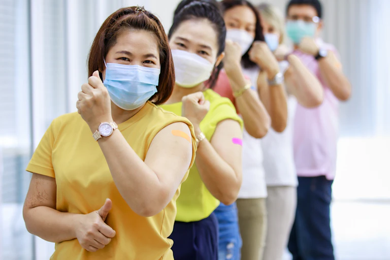 Five people lined up wearing facemasks and flexing their arm with a bandaid on the bicep