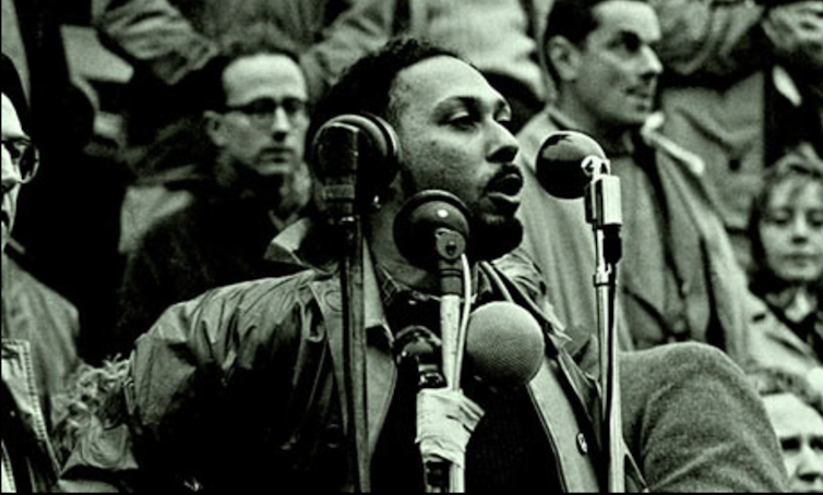 A young bearded black man addresses a crowd at a microphone.