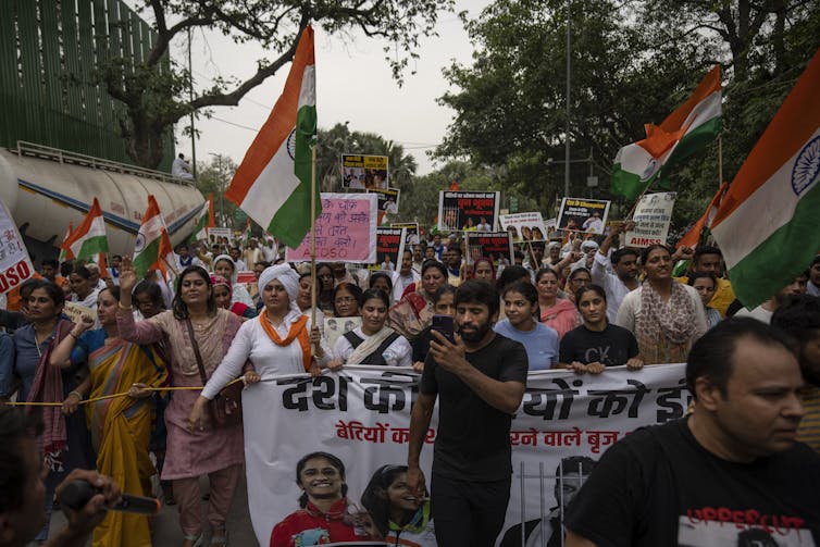 A man in black at a protest holding his phone. A group of people march behind him holding signs and waving flags.