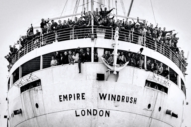 A large white ship called Empire Windrush carrying migrants from the Caribbean to London, with people on deck waving and cheering.