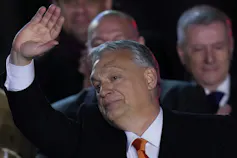 A grey-haired round-faced man in a suit waves.
