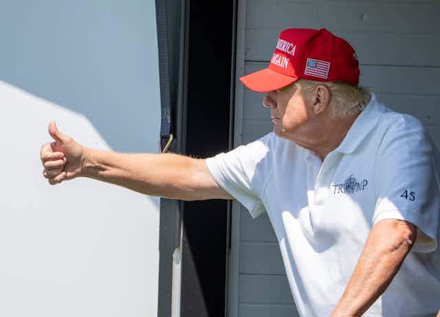 An older rotund man in a white T-shirt and red ball cap gives the thumbs up.