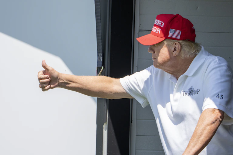 An older rotund man in a white T-shirt and red ball cap gives the thumbs up.