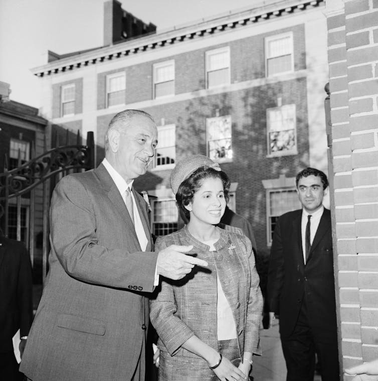 A white man dressed in a business suit talks with a Black woman who is smiling.