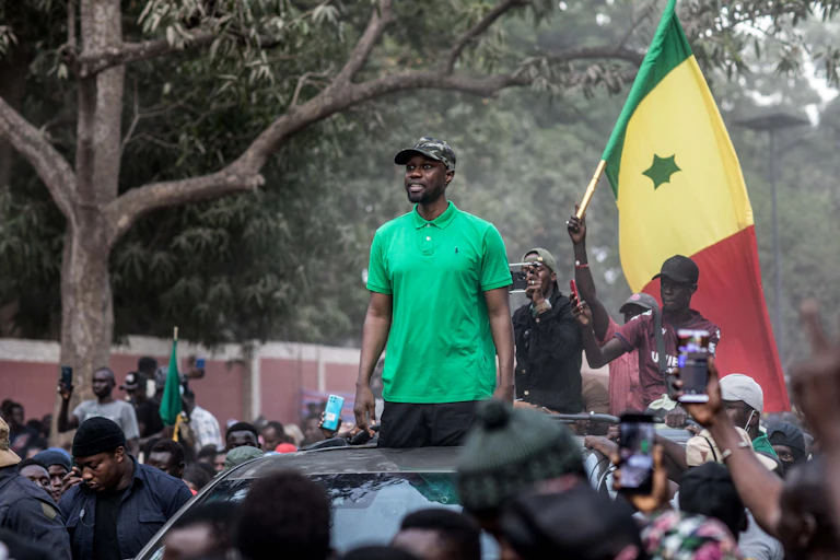 Un homme arborant une chemise verte et une casquette, debout dans une voiture décapotable, au milieu d'une foule.