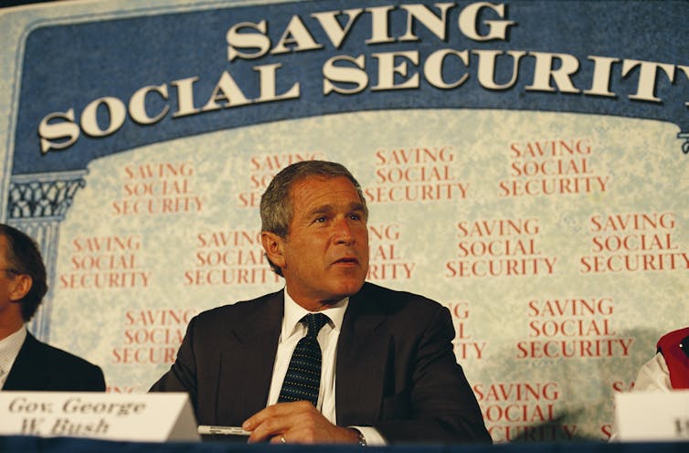 A man with gray hair sits at a table in front of a giant replica of a Social Security card.