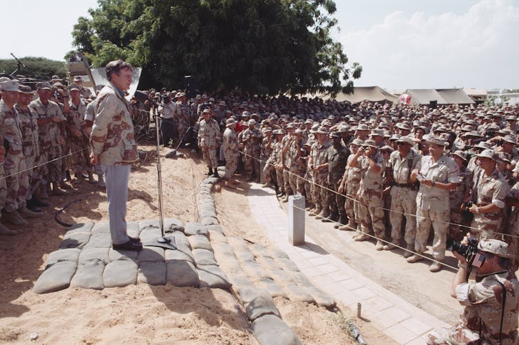 A white man gives a speech while standing near hundreds of American soldiers.