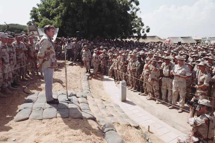 A white man gives a speech while standing near hundreds of American soldiers.