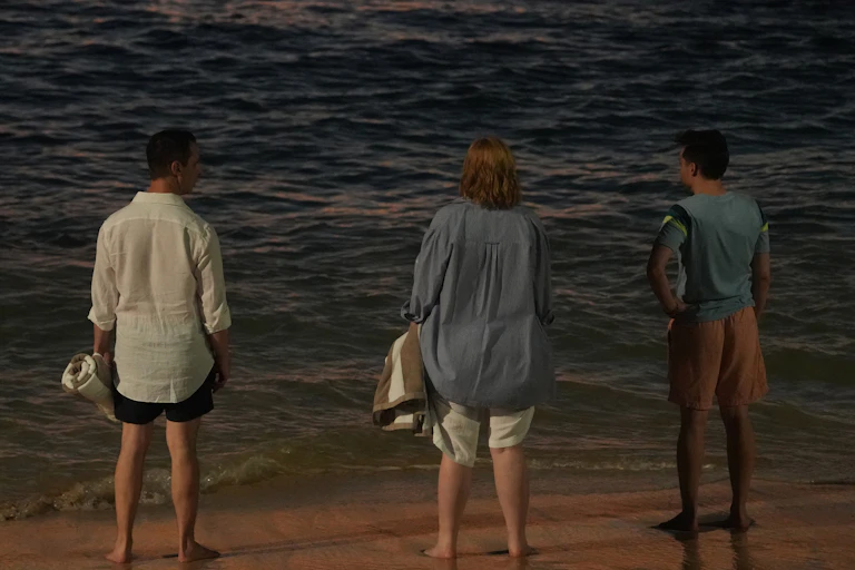 Jeremy Strong, Sarah Snook and Kieran Culkin stand on a beach looking out to sea, shot from behind.