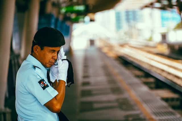 Thai security guard at a station in Bangkok taking off his mask to wipe sweat from his face