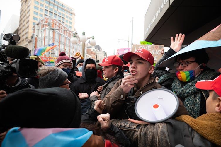 A man with a megaphone in the middle of a chaotic crowd of people. Rainbow flags are seen in the background.