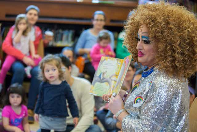 A drag queen in a silver dress reads to young children sitting with their parents.