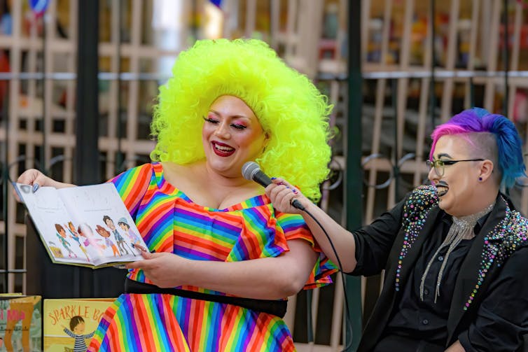 A drag queen in a multi-coloured dress with bright yellow hair reads from a children's book.