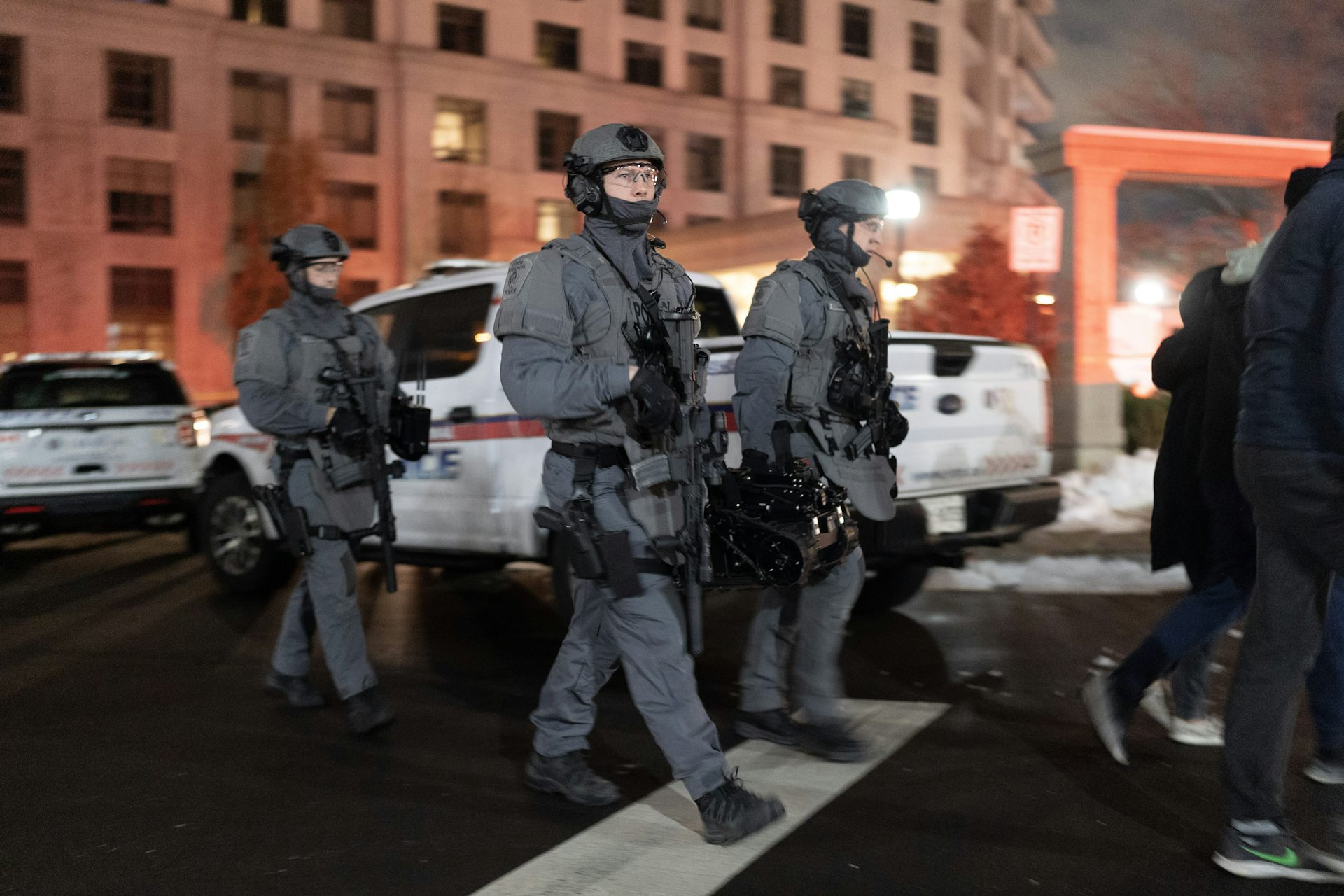 Police in grey tactical uniforms with helmets and face masks carry assault rifles.