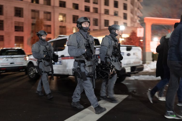 Police in grey tactical uniforms with helmets and face masks carry assault rifles.