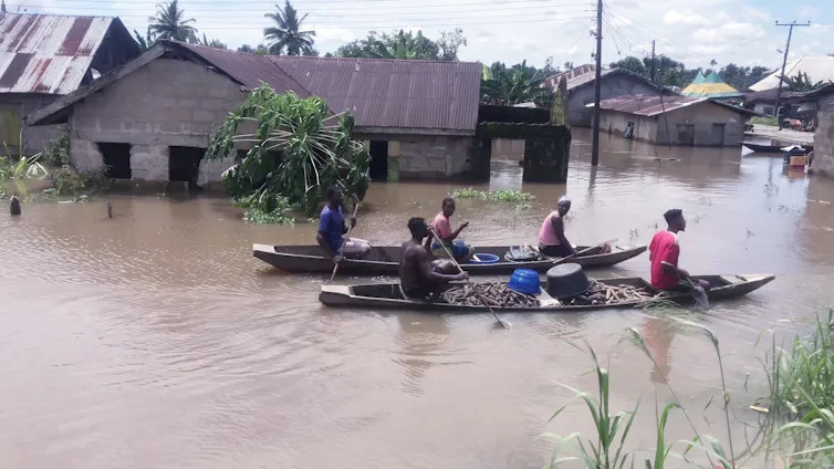 People paddle canoes down a flooded residential street past half-submerged residential houses.