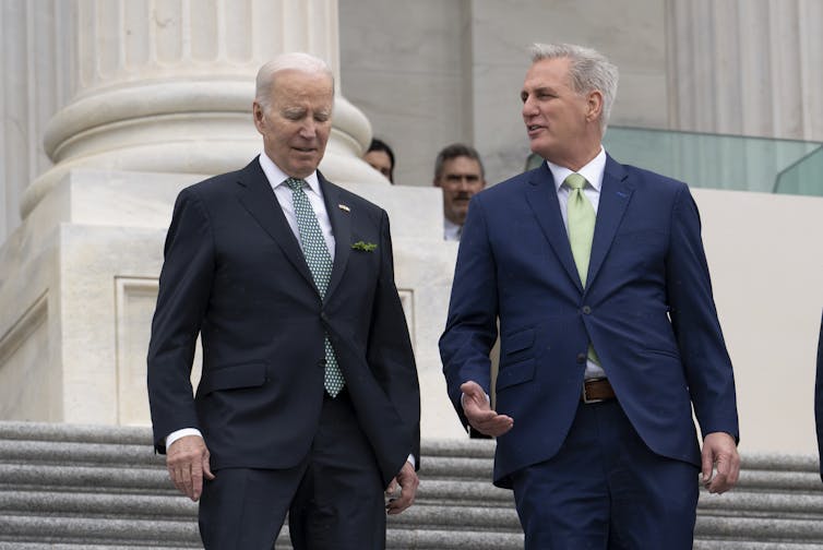 Two gray-haired men in dark suits and white shirts and ties, standing outside on a large set of steps.