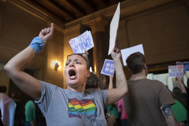 A person with a rainbow on their shirt holds up a hand with a pointed finger and a sign in the other hand. They appear to be yelling.