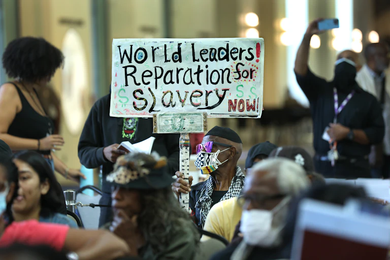 A man is holding up a sign in support of reparations for formerly enslaved people during a crowded hearing.