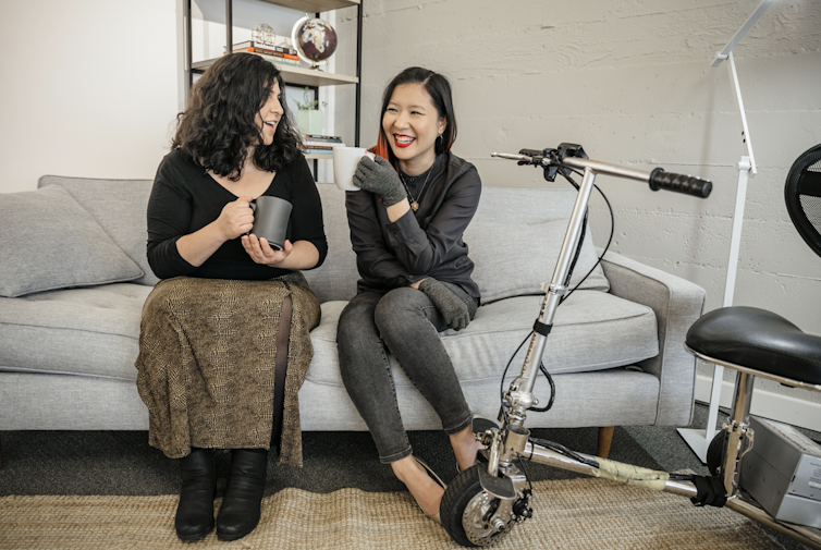 Latinx disabled woman and an Asian disabled genderfluid person chat and sit on couch, both holding coffee mugs. Electric lightweight mobility scooter rests on the side