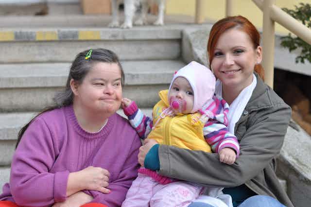 Woman with Down's syndrome sitting on outside step with other woman, young child on lap