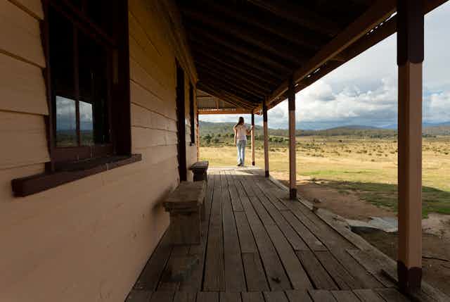 Woman stands on veranda of weatherboard house looking at hills and countryside