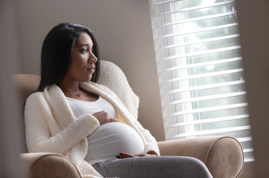 A pregnant woman holds her stomach while looking out the window of her home.