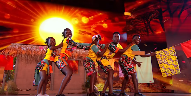 Five children in colourful African clothing stand in a dance line with their hands on the shoulders of the child in front of them, on a stage decorated with a sunset background, small hut and towels hanging on a laundry line