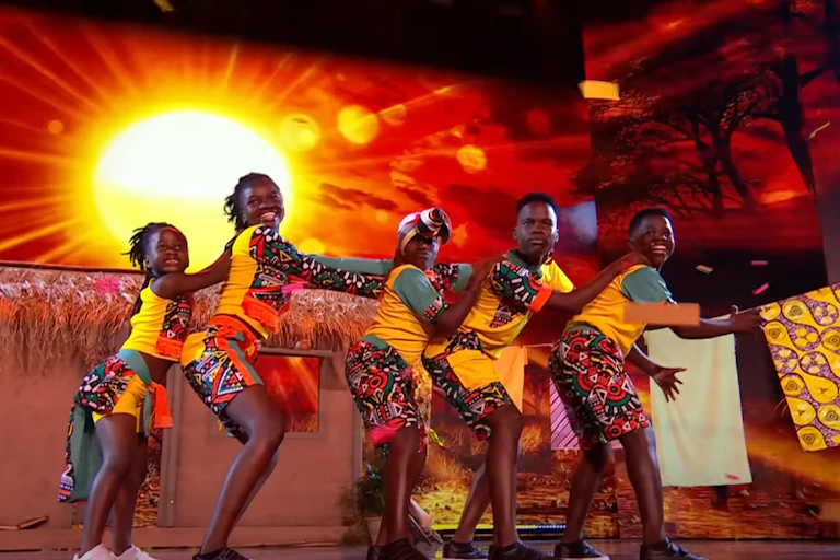 Five children in colourful African clothing stand in a dance line with their hands on the shoulders of the child in front of them, on a stage decorated with a sunset background, small hut and towels hanging on a laundry line