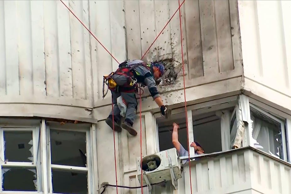 A man hangs from a harness examining damage to an apartment building after a drone attack.