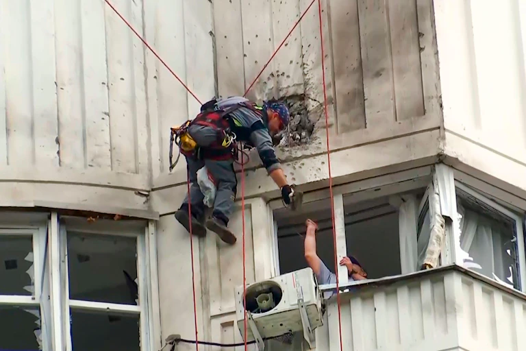 A man hangs from a harness examining damage to an apartment building after a drone attack.