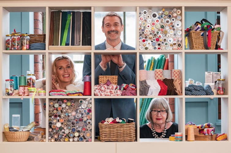 Great British Sewing Bee presenters, Sara Pascoe, Patrick Grant and Esme Young posing behind shelves of sewing materials.