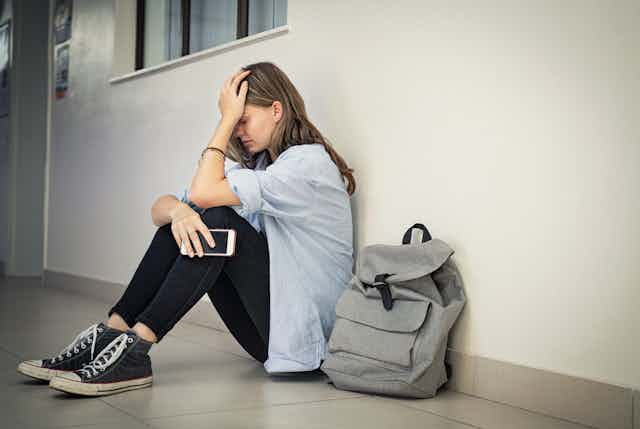 Una niña sentada al lado de su mochila llora, con el teléfono en la mano.