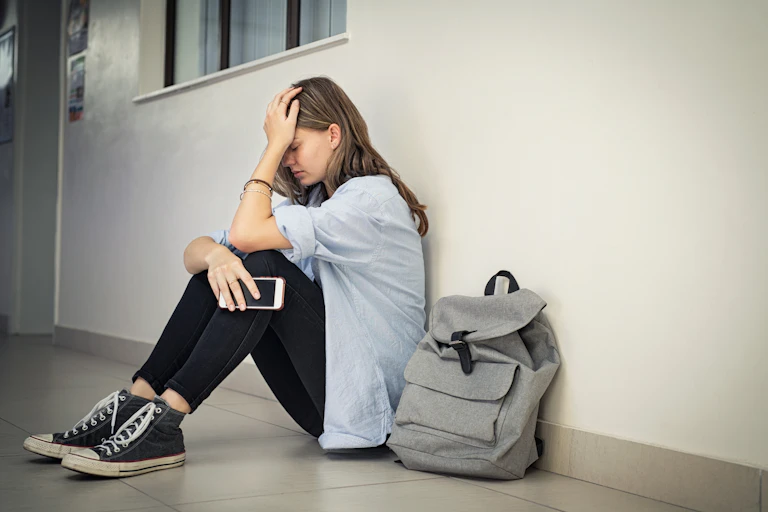 Una niña sentada al lado de su mochila llora, con el teléfono en la mano.