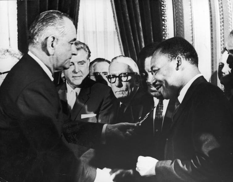 A middle-aged white man stands in front of a crowd of people as he hands a pen to a Black man dressed in a business suit.