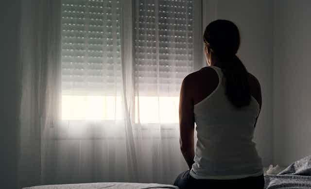 A woman sits on a bed staring out the window in a dark room.