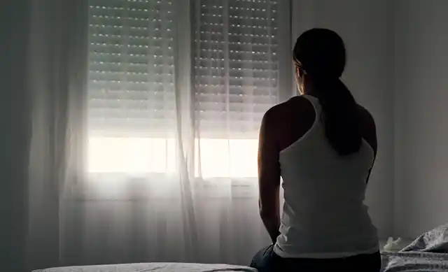 A woman sits on a bed staring out the window in a dark room.