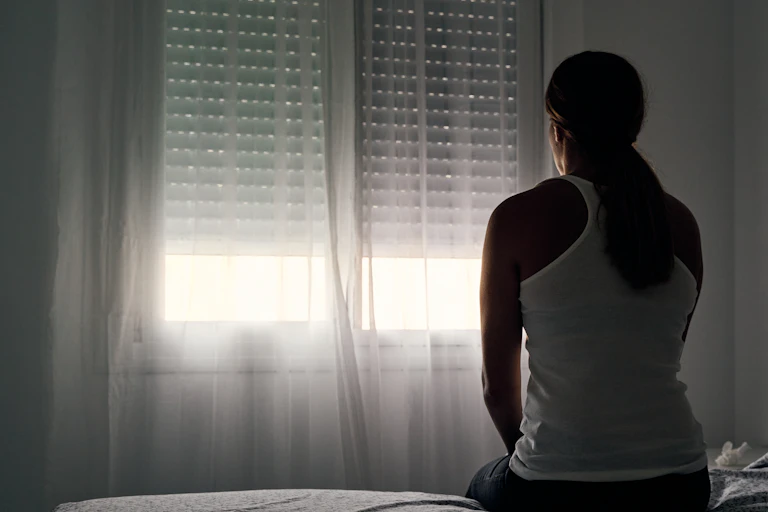 A woman sits on a bed staring out the window in a dark room.