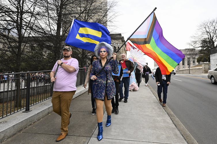 A group of people walk down a sidewalk carrying flags promoting equality and LGBTQ+ rights