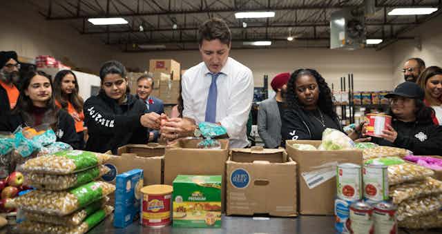 A dark-haired man in a white shirt and tie packs boxes with food surrounded by food bank workers.
