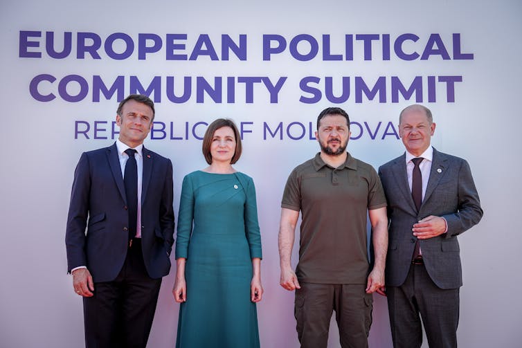 Two men in suits stand in a row with a woman in a teal dress and a man wearing camouflage green in front of a sign that says European Political Community Summit.
