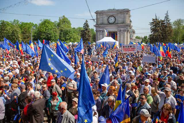 A large group of people appear to take part in a large protest and wave blue flags with little yellow stars. It is a clear, sunny day and there are trees in the background and a large tower that has a clock on it. 