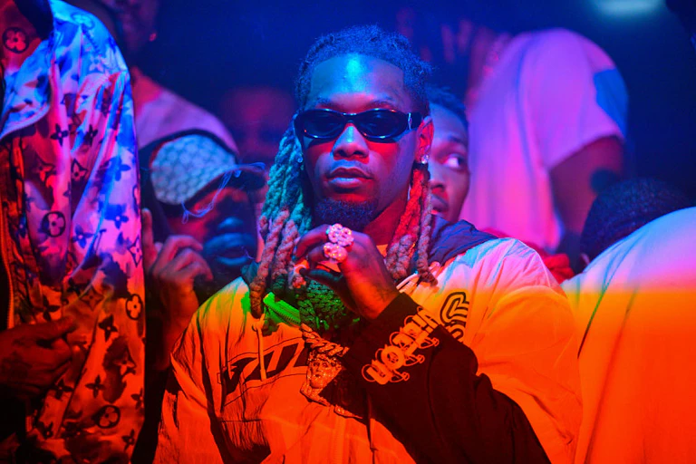A man in dark sunglasses is bathed in multicolored lights.