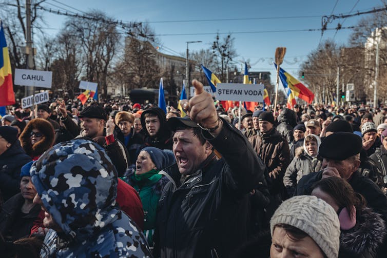 A large group of people appear to attend a protest, with one person raising his hand in the air and people hold red, blue and yellow striped flags.