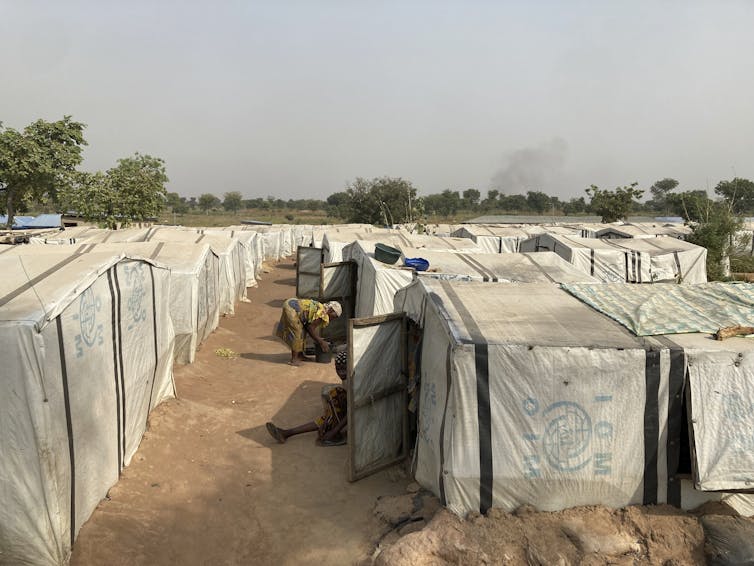 People sit outside square white tents in the middle of a dirt-packed field.