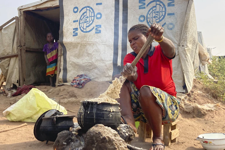 A Nigerian woman stirs a flour mixture in a cast iron post outside of a white temporary shelter. A young girl stands in the entryway of the shelter.