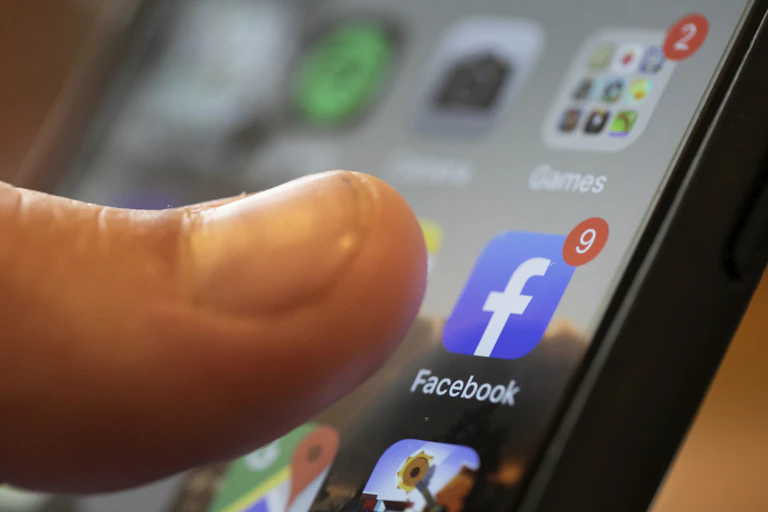 close-up of a smart phone home screen with a thumb poised over a blue icon with a white letter f