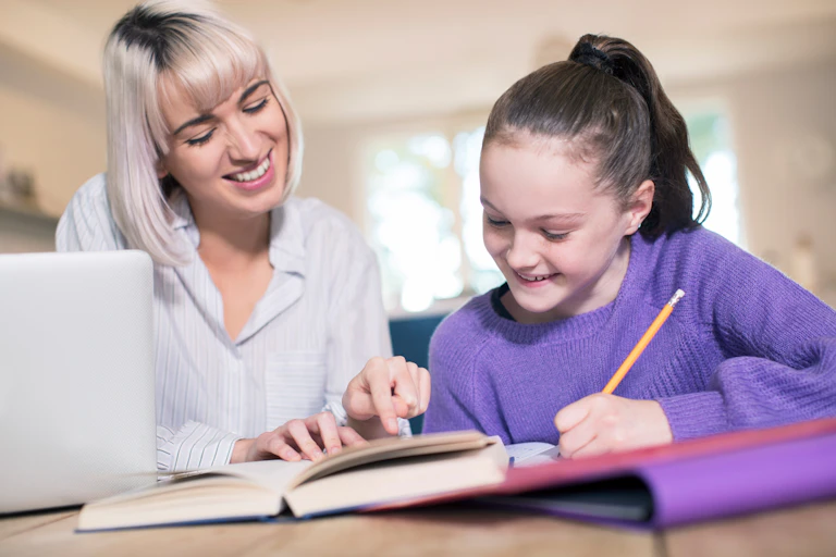 A tutor and young girl sit at a desk, with books and laptop.